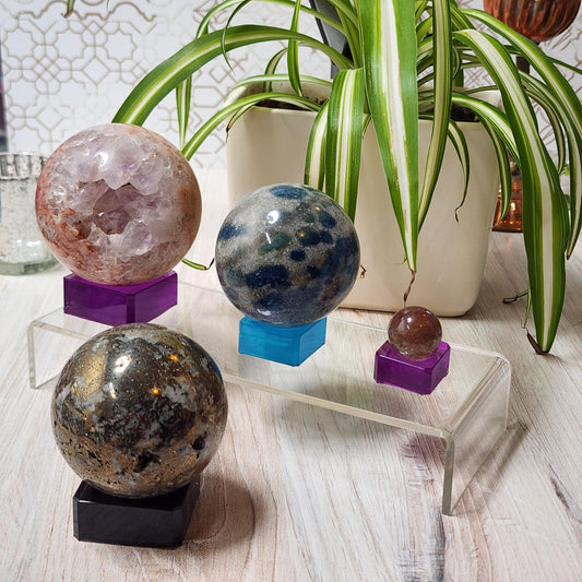 A decorative arrangement featuring four colorful crystal spheres in various sizes, each gracefully perched on The Crystalary's Custom-made Crystal Sphere Holders, designed for endless color combinations. These sit atop a clear, elevated stand with a light wood surface beneath. In the background, a potted spider plant with green and white striped leaves adds a natural touch.