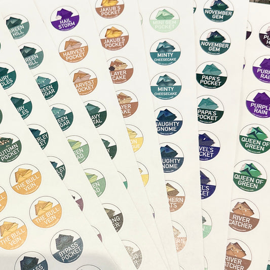 Fan-like arrangements of full-color circular stickers, each labeled with distinctive names such as "HAIL STORM," "PAPA'S POCKET," and "NAUGHTY GNOME." These stickers, part of The Crystalary's Diana Maria Passport Stickers collection, showcase a variety of colors and mountain-like icons behind the text.