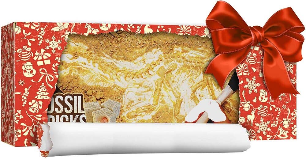 A rectangular gift box adorned with festive holiday wrapping and topped with a red bow contains the Dig a Dozen Fossil Bricks kit by Dan&Darci. Inside, beige dig bricks, a small chisel, and a brush are partially unwrapped to reveal unique fossils. The packaging features holiday-themed designs and illustrations.