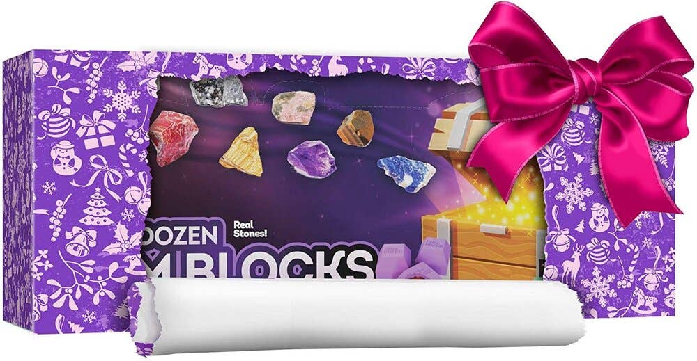 A gift box with purple, festive wrapping featuring white snowflakes, Christmas trees, and gingerbread men is partially unwrapped to reveal a set of twelve colorful gemstones labeled "Dig a Dozen Gem Blocks" by Dan&Darci. A pink ribbon and bow adorn the box. Gem learning cards inside help you identify these unique real gems.