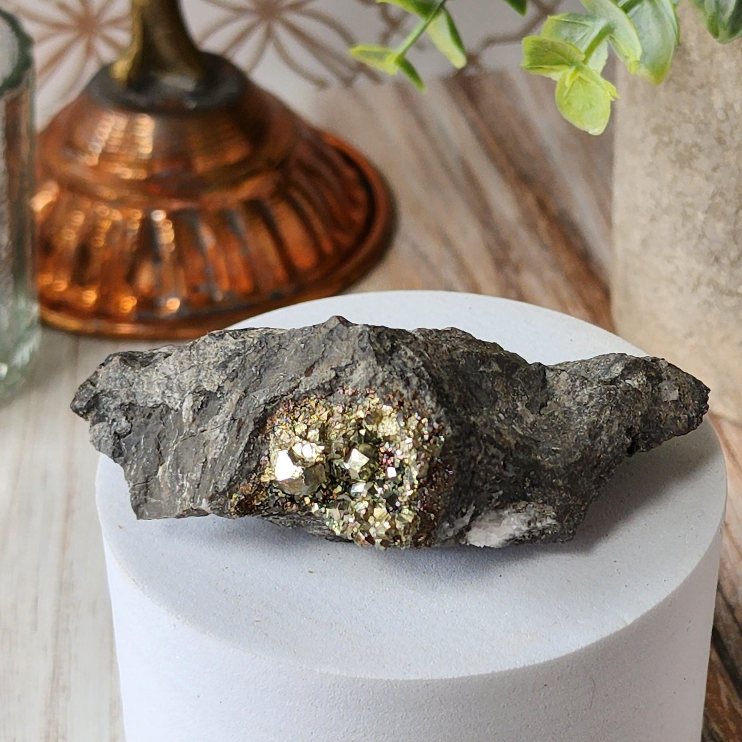 A piece of Duff Quarry Pyrite in Silurian dolostone from The Crystalary, with embedded shiny metallic crystals, is elegantly displayed on a round white pedestal. A copper container, a glass jar, and a partially visible green plant adorn the background, creating an indoor setting with a decorative ambiance.