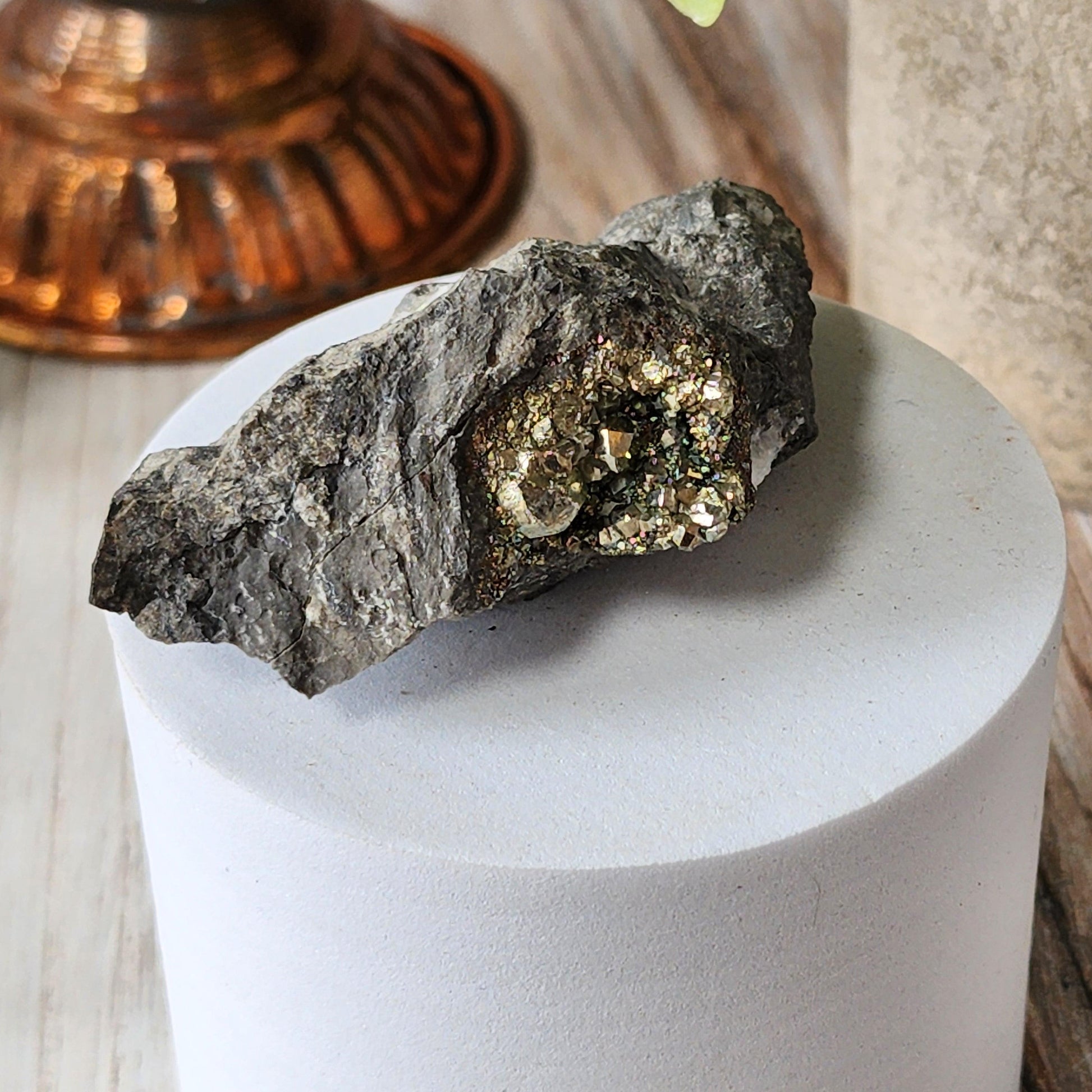 The Crystalary's Duff Quarry Pyrite in Silurian dolostone is showcased on a white cylindrical platform, featuring a cluster of shiny, metallic pyrite crystals. In the background, there’s a slightly out-of-focus copper-colored object and part of a textured vase.