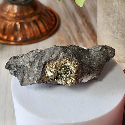 A piece of Duff Quarry Pyrite in Silurian dolostone from The Crystalary, featuring a cluster of shiny, gold-colored pyrite crystals embedded in rough gray rock, is placed on a round, white pedestal. In the background, there is a copper-toned decorative object and a glimpse of a green leaf.