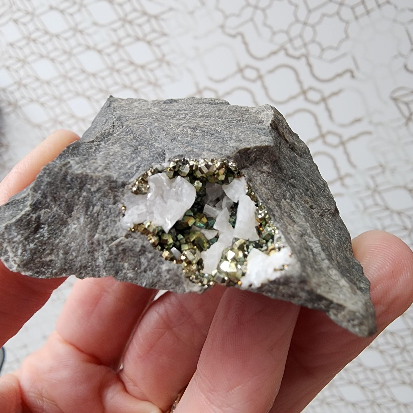 A close-up photo of a hand holding the Duff Quarry Pyrite in Silurian dolostone from The Crystalary, showcasing its cavity filled with shiny pyrite crystals and white quartz formations. The background features a light-colored, intricate geometric pattern, creating a beautiful contrast with the diploid crystal formation within the rock.