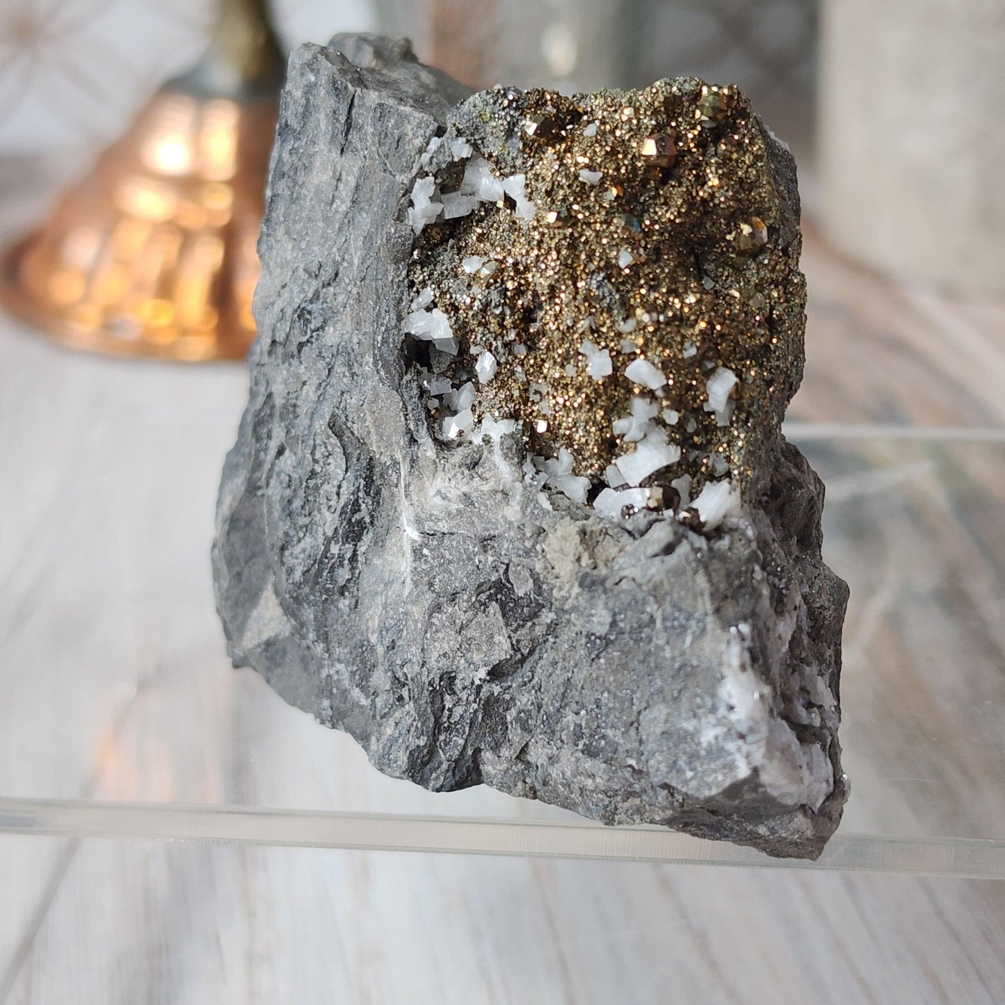 A close-up photo of a Duff Quarry Pyrite in Silurian dolostone by The Crystalary showcases a cluster of shiny, golden pyrite crystals and white quartz formations embedded in it. The rock rests on a clear surface with a blurred background featuring warm hues and subtle, out-of-focus details.