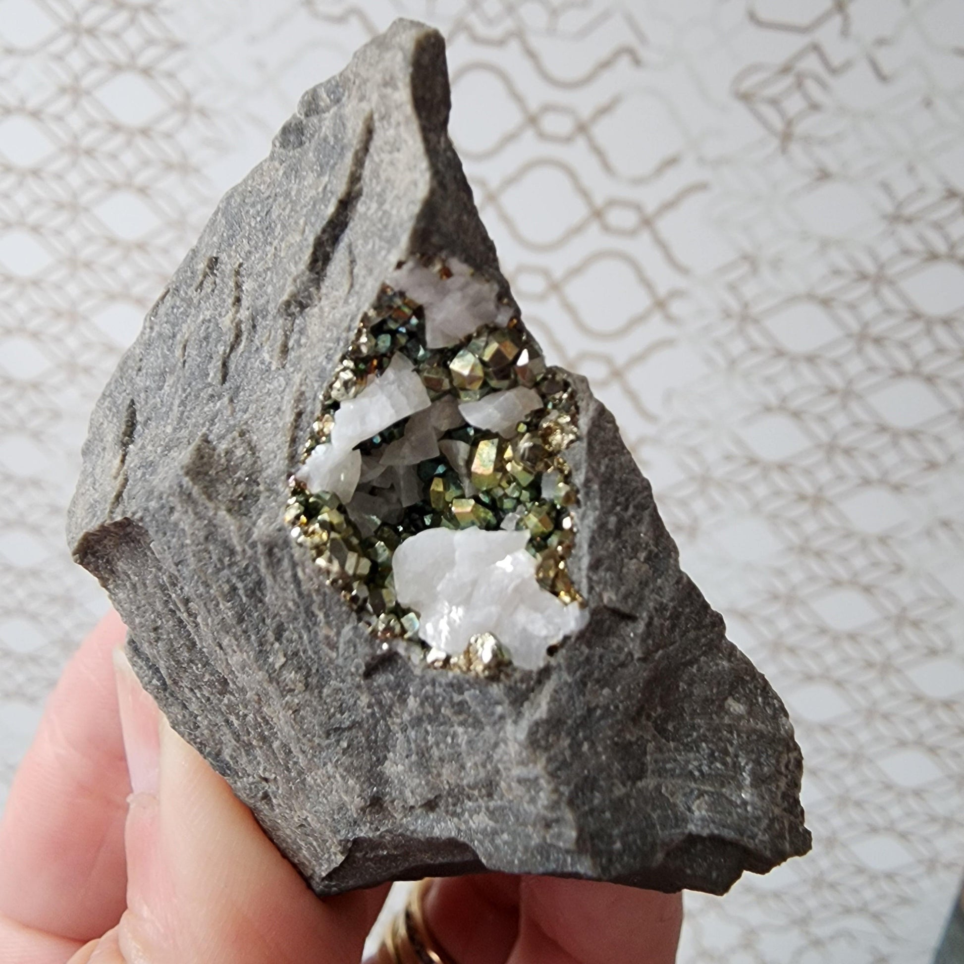 A hand is holding a specimen of Duff Quarry Pyrite in Silurian dolostone by The Crystalary; the gray rock boasts a rough exterior, while inside, shiny white crystals are surrounded by small, reflective pyrite minerals. This stunning piece stands out against the light-colored pattern with interlocking shapes in the background.