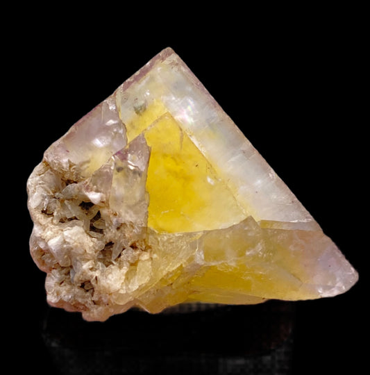 A vibrant yellow Fluorite- Annabel Lee Mine from Harris Creek, Hardin County, Illinois, USA, by The Crystalary sits on a black background. The triangular crystal has sharp, translucent facets with a cluster of rough, whitish material attached to one side, providing a striking contrast to the smooth surfaces. Natural imperfections and internal textures are visible.