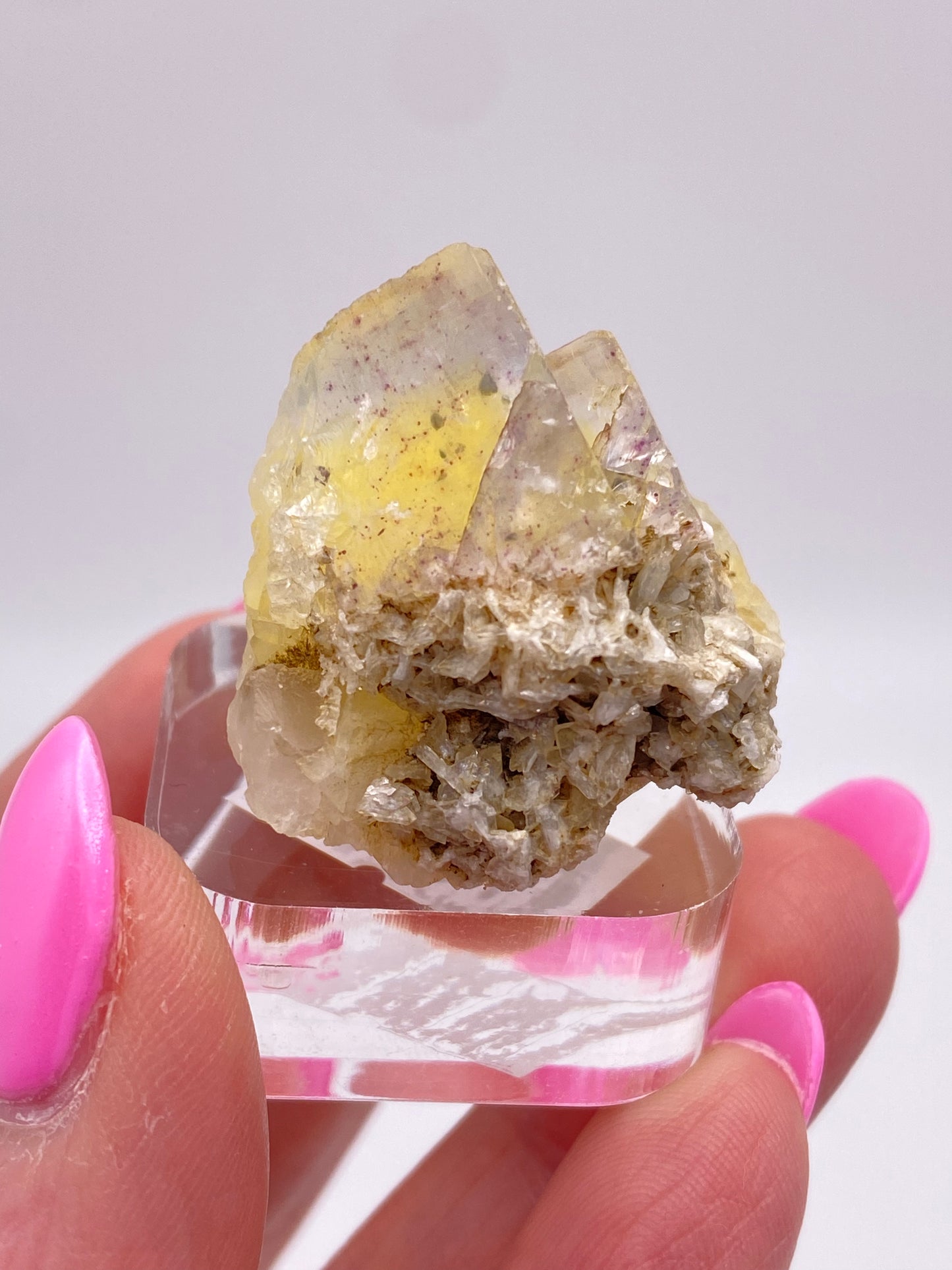 A hand with bright pink nails holds a small cluster of yellow fluorite from the Annabel Lee Mine, Harris Creek, Hardin County, Illinois, USA on a clear base. This lovely piece from The Crystalary features pale yellow fluorite crystals that are slightly translucent with rough, textured surfaces and some embedded mineral deposits. The background is white and out of focus.