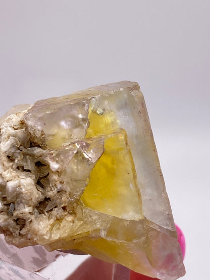 Close-up of a translucent yellow Fluorite- Annabel Lee Mine, Harris Creek, Hardin County, Illinois, USA crystal from The Crystalary with a pale yellow hue. The crystal features a jagged surface and some white encrustations, showcasing rough and smooth textures. Sourced from the Annabel Lee Mine in Hardin County, the background is plain and out of focus.