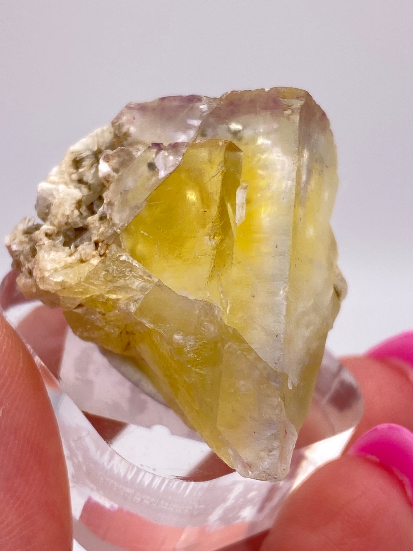 A close-up image features a hand with pink nail polish holding a translucent yellow fluorite crystal, sourced from the Annabel Lee Mine in Harris Creek, Hardin County, Illinois. This product from The Crystalary shows visible inclusions and imperfections and is partially embedded in a light-colored rock matrix. The background is out of focus.