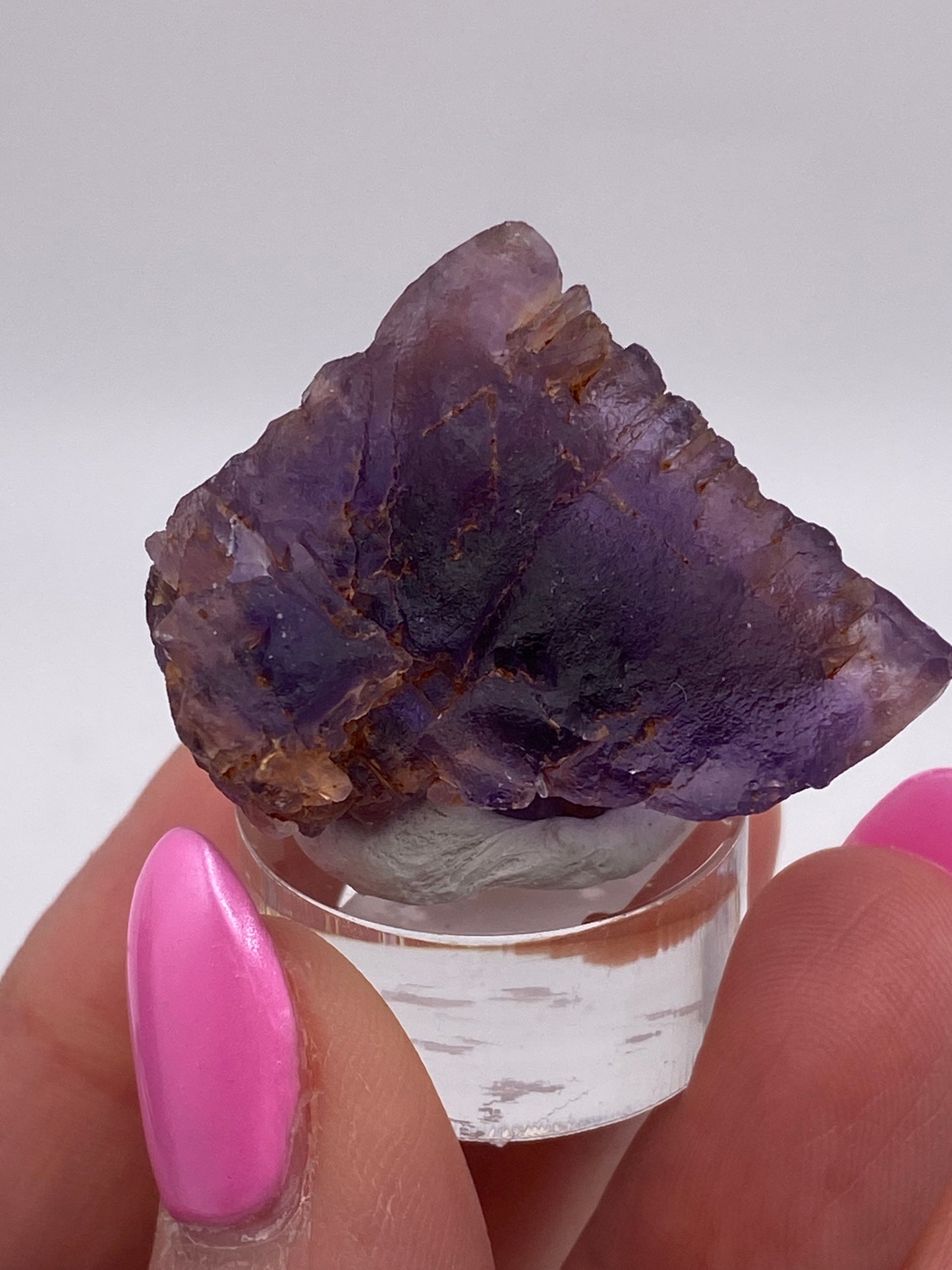 A person with bright pink, manicured nails holds a roughly hewn "Fluorite- Cave in Rock, Hardin County, Illinois, USA" crystal from The Crystalary on a small round clear stand against a plain light-colored background.