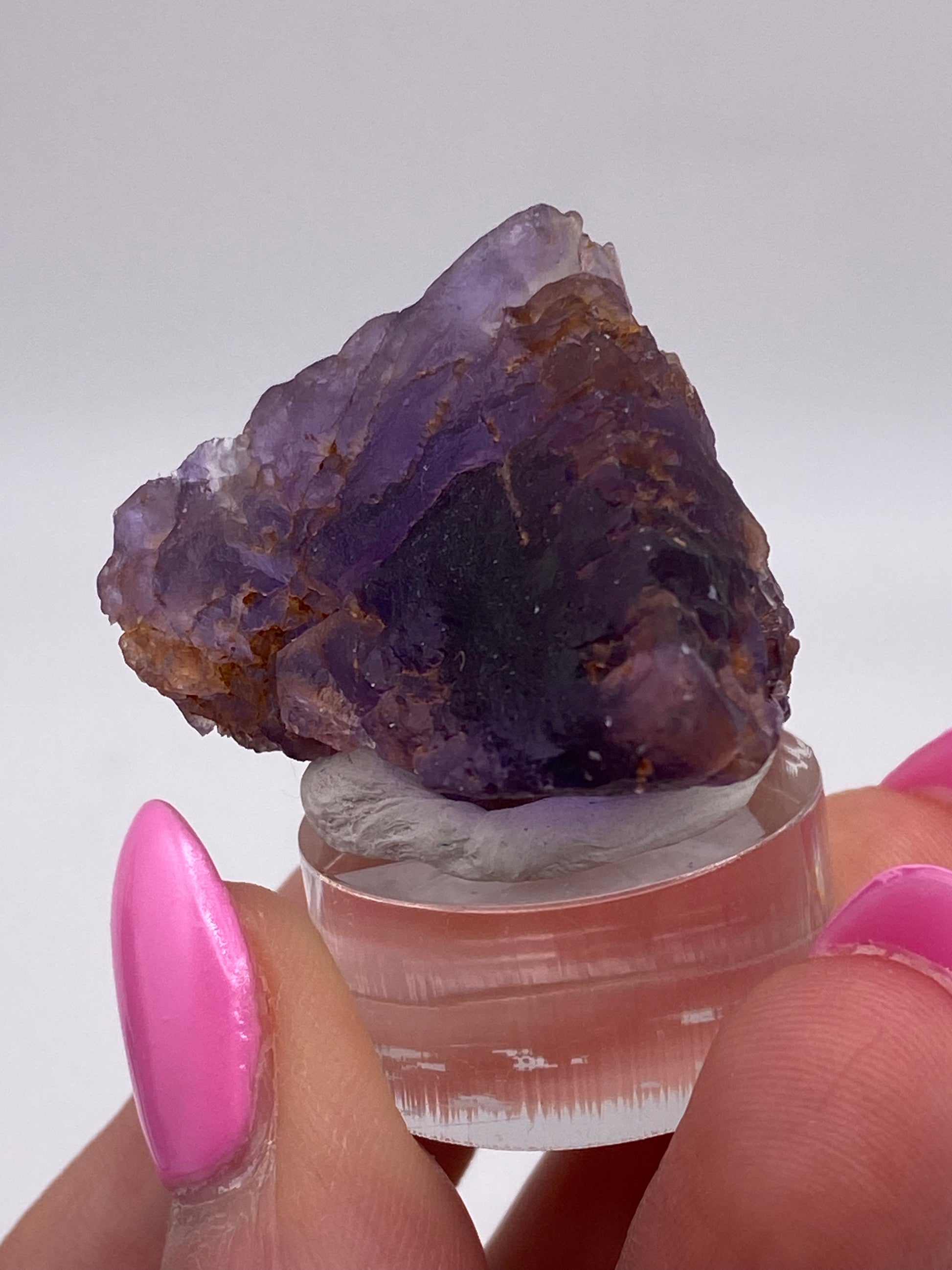 A hand with bright pink nails holds a small, rough Fluorite crystal from Cave in Rock, Hardin County, Illinois, USA, secured on a clear plastic stand. The crystal is primarily purple with some brownish areas, highlighting its raw and unpolished nature. The background is plain and white. This captivating piece is brought to you by The Crystalary.