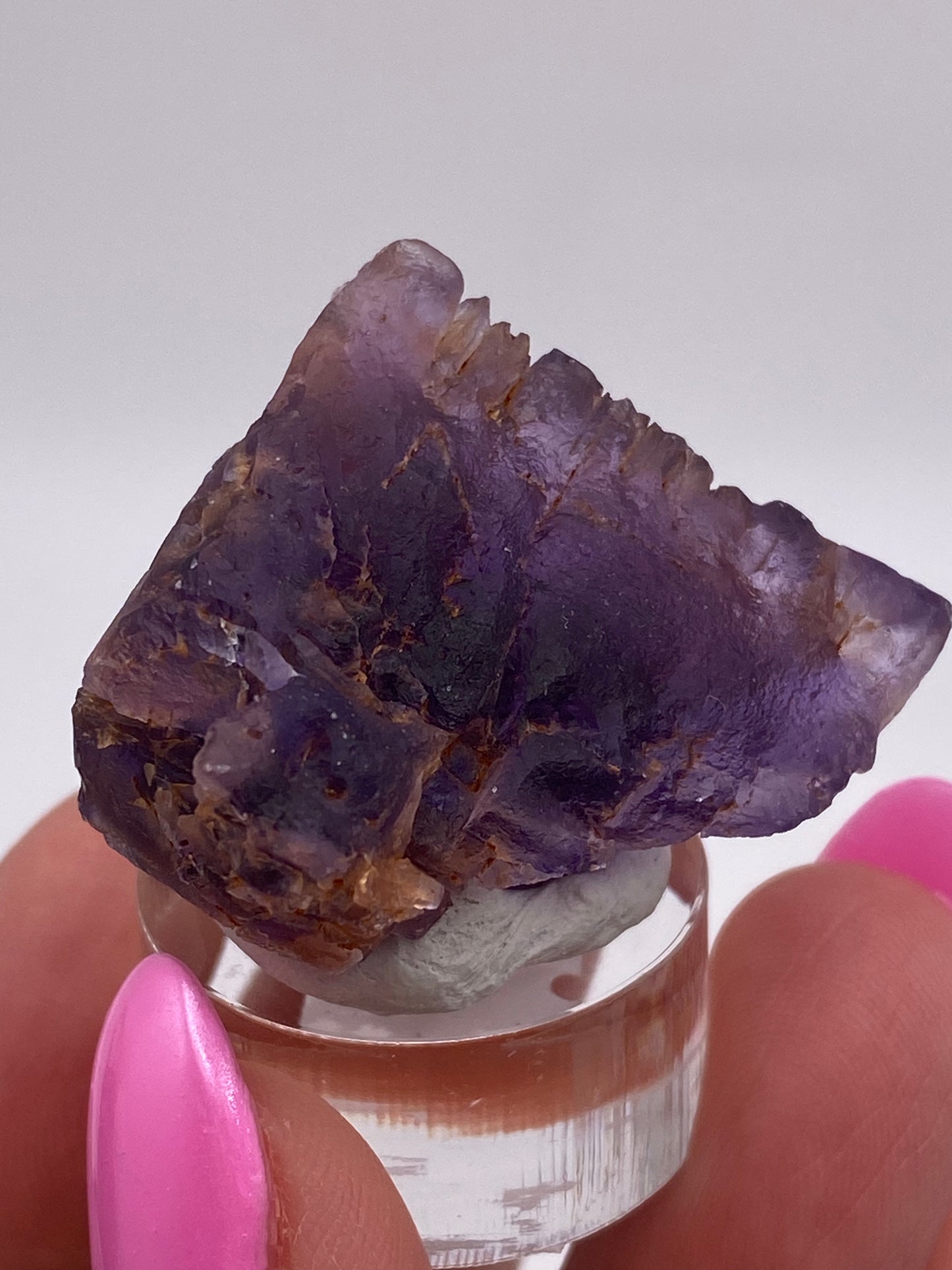 A close-up shot features a person with pink nails holding a small, rough piece of Fluorite from Cave in Rock, Hardin County, Illinois, USA by The Crystalary. This gemstone has partially translucent qualities and exhibits a mix of deep purple and brownish hues with natural cleavages and irregular edges. It is displayed on a clear stand.