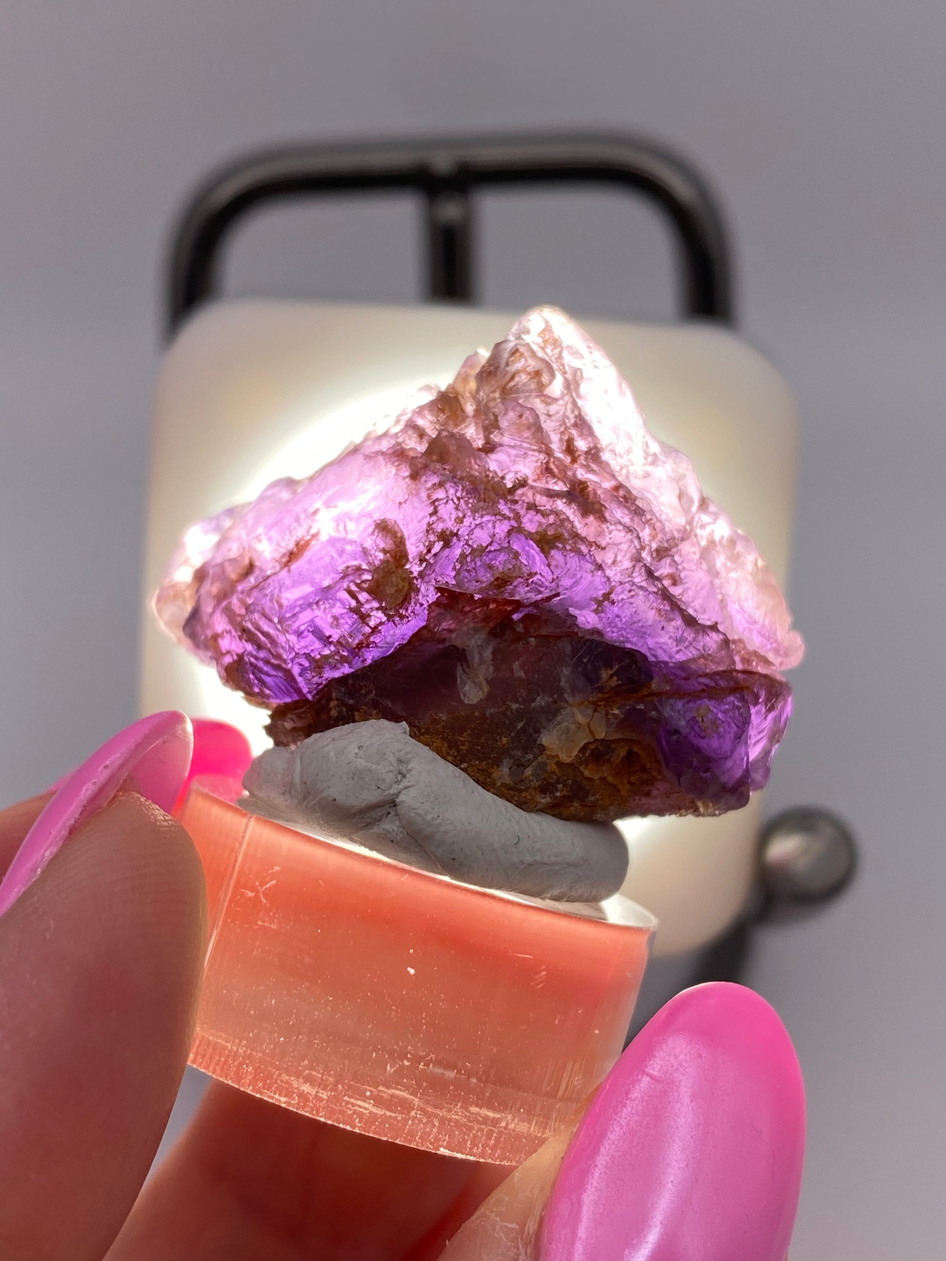 Close-up of a person holding a piece of raw Fluorite- Cave in Rock, Hardin County, Illinois, USA by The Crystalary. The gemstone is positioned on a small, round stand and illuminated from behind, highlighting its vibrant purple hue. The person has pink-painted nails. This stunning specimen originates from the renowned Hardin County, Illinois fluorite mines.