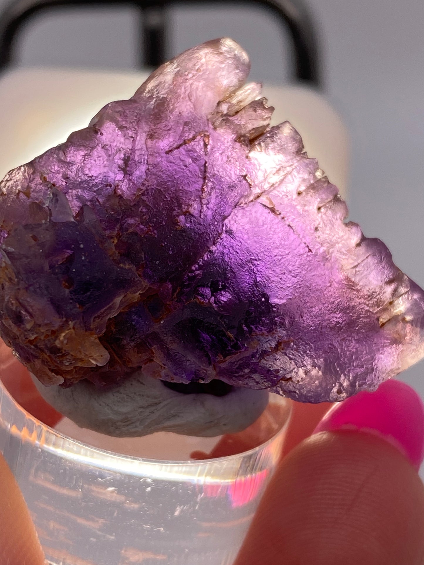 A close-up photograph features a person's hand holding a vibrant Fluorite- Cave in Rock, Hardin County, Illinois, USA crystal from The Crystalary against a blurred background. The crystal showcases rich, deep purple hues and has a jagged, rough surface texture. The person's fingernail is painted pink.