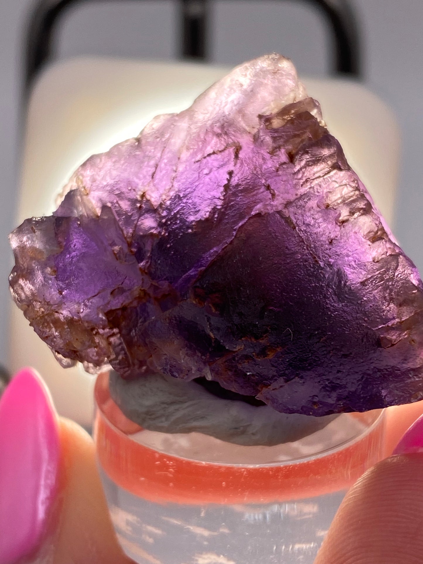 Close-up of "Fluorite- Cave in Rock, Hardin County, Illinois, USA" by The Crystalary held between two fingers with bright pink nail polish. The purple fluorite crystal is roughly textured and backlit, revealing its translucent qualities and deep violet hues. It rests on a small, clear stand.