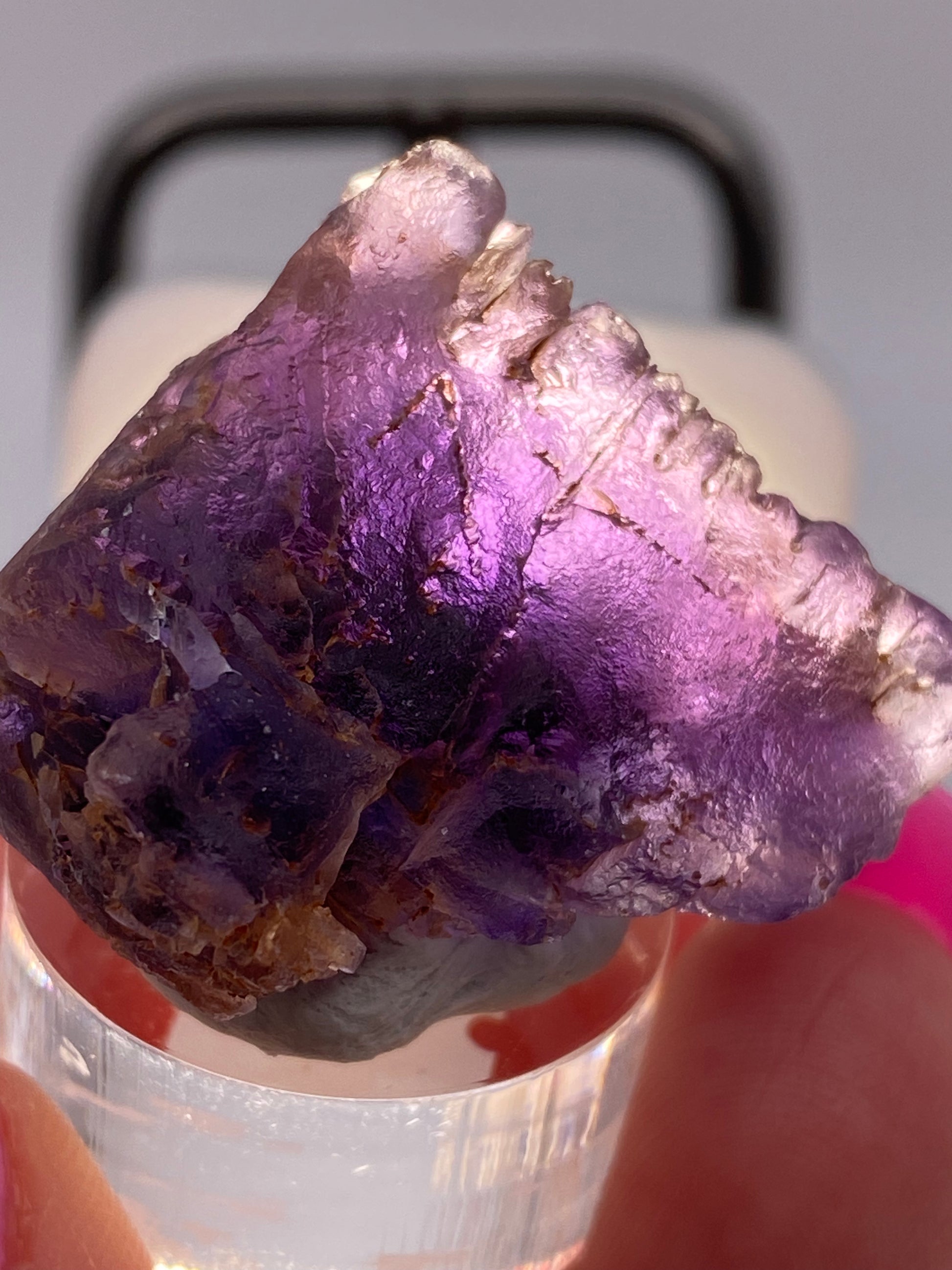 A close-up of a raw fluorite crystal from Cave in Rock, Hardin County, Illinois, being held in a person's hand. The crystal, offered by The Crystalary, features a deep purple color with rough, jagged edges and is partially translucent. Light shines through the crystal to enhance its vivid hues, reminiscent of the striking purple amethyst. The background is softly blurred.