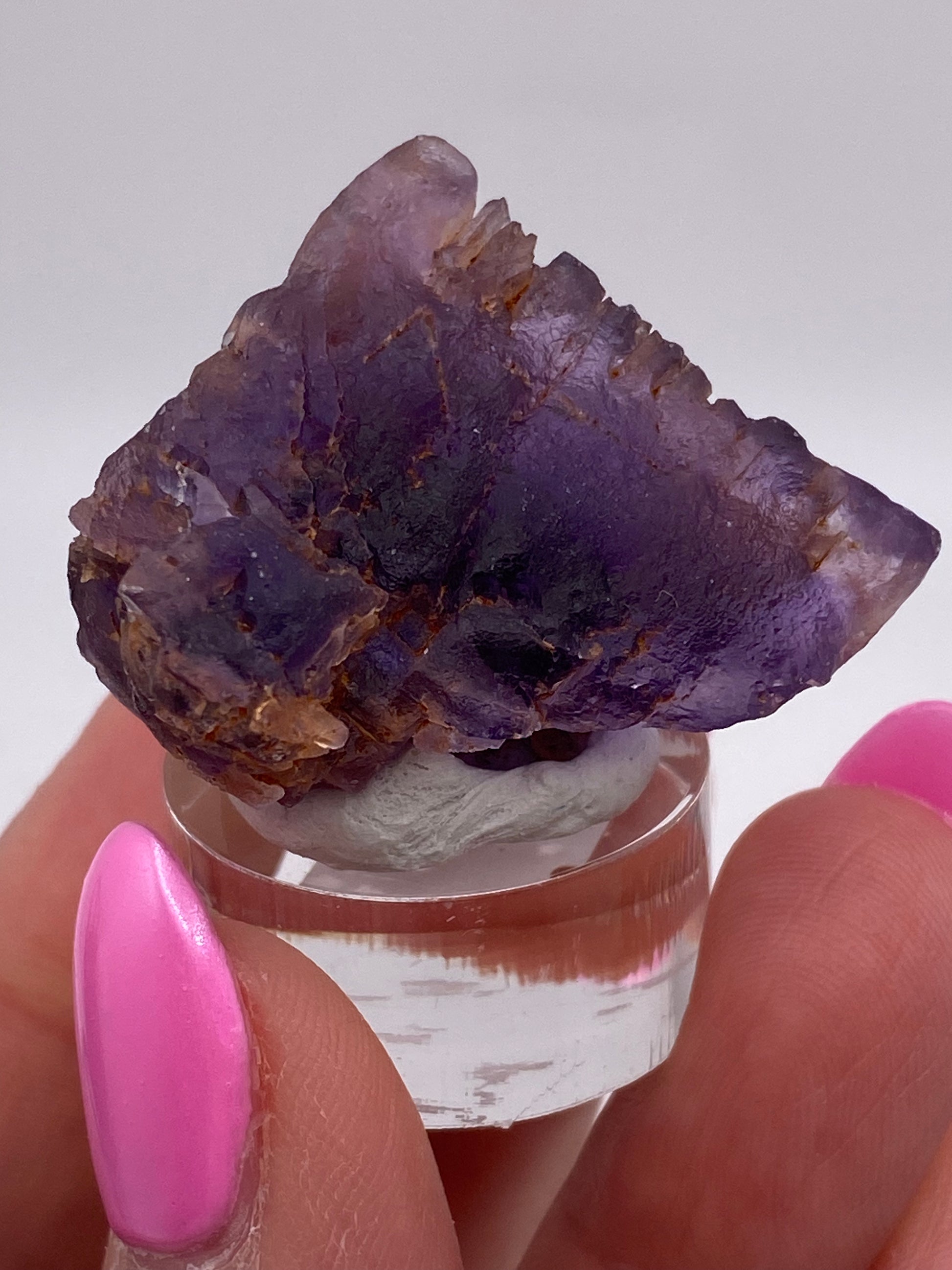 A close-up of a person’s hand with pink-painted nails holding a small, translucent purple specimen from The Crystalary's Fluorite collection, sourced directly from Cave in Rock, Hardin County, Illinois. The fluorite rests on a clear display stand and features a rough, jagged texture with deep violet hues and some brown inclusions.