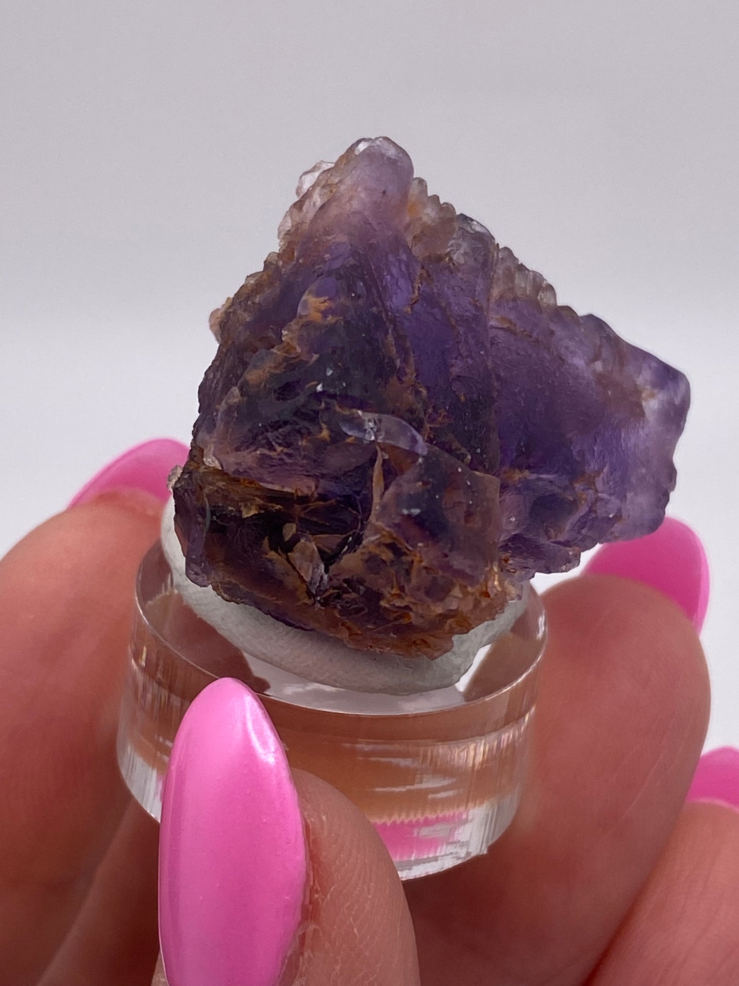 A person with pink-painted nails holds a small, deep purple amethyst crystal, mounted on a round, clear stand. The background is plain and out of focus, drawing attention to the intricate details and rich color reminiscent of The Crystalary's Fluorite from Cave in Rock, Hardin County, Illinois.