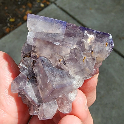 A hand holding a large, light purple fluorite crystal cluster with visible geometric formations. The glassy cluster is semi-transparent and has a reflective surface, catching glimmers of light. The background is a concrete walkway, slightly out of focus. This stunning specimen from "The Crystalary" hails from Cromwell Pocket, Greenlaws Mine in Daddry Shield, Stanhope, County Durham, England.