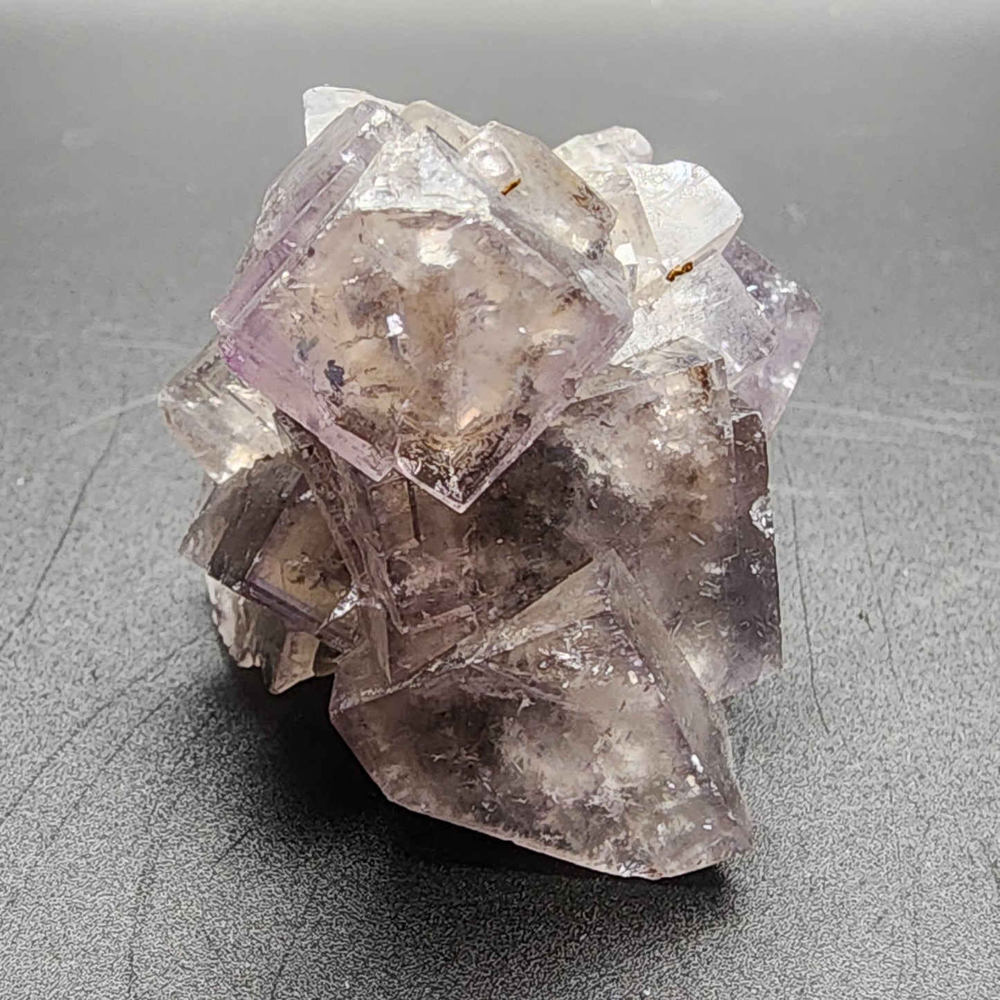 A glassy cluster of purple and clear cubic crystals from The Crystalary’s Fluorite collection, sourced from the Cromwell Pocket in Greenlaws Mine, Stanhope, County Durham, sits on a smooth dark gray surface. The semi-transparent fluorite crystals feature various inclusions and fractures within them, creating an intricate and textured appearance that seems to change color with the light.