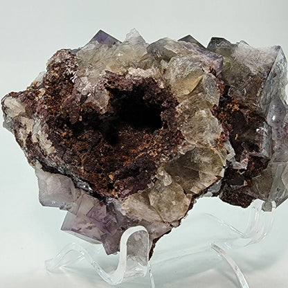 A rugged, multi-faceted Fluorite specimen from Cromwell's Pocket at the Greenlaws Mine in County Durham, England. It features a mix of translucent and opaque surfaces, with a darker and more porous central area that may indicate different mineral compositions. This exquisite piece from The Crystalary rests on a clear display stand against a plain background.