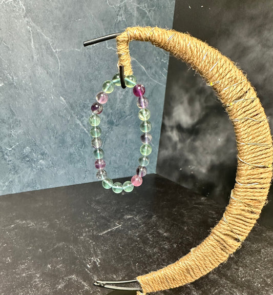 The Crystalary's Fluorite Crystal Bracelet, featuring alternating light green and purple beads, is elegantly displayed on a crescent-shaped stand wrapped in twine. This stand, ideal for showcasing gemstone bracelets, is set against a backdrop with a marbled pattern on the left and a dark, smoky texture on the right.