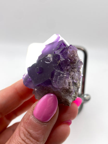 A close-up of a hand with pink-painted nails holding a rough piece of Fluorite, ex. Ben Clement- Cave in Rock, Hardin County, Illinois from The Crystalary. The mineral exhibits a combination of crystalline and granular textures, showcasing its vibrant violet hues. The background is blurred, focusing attention on the fluorite.