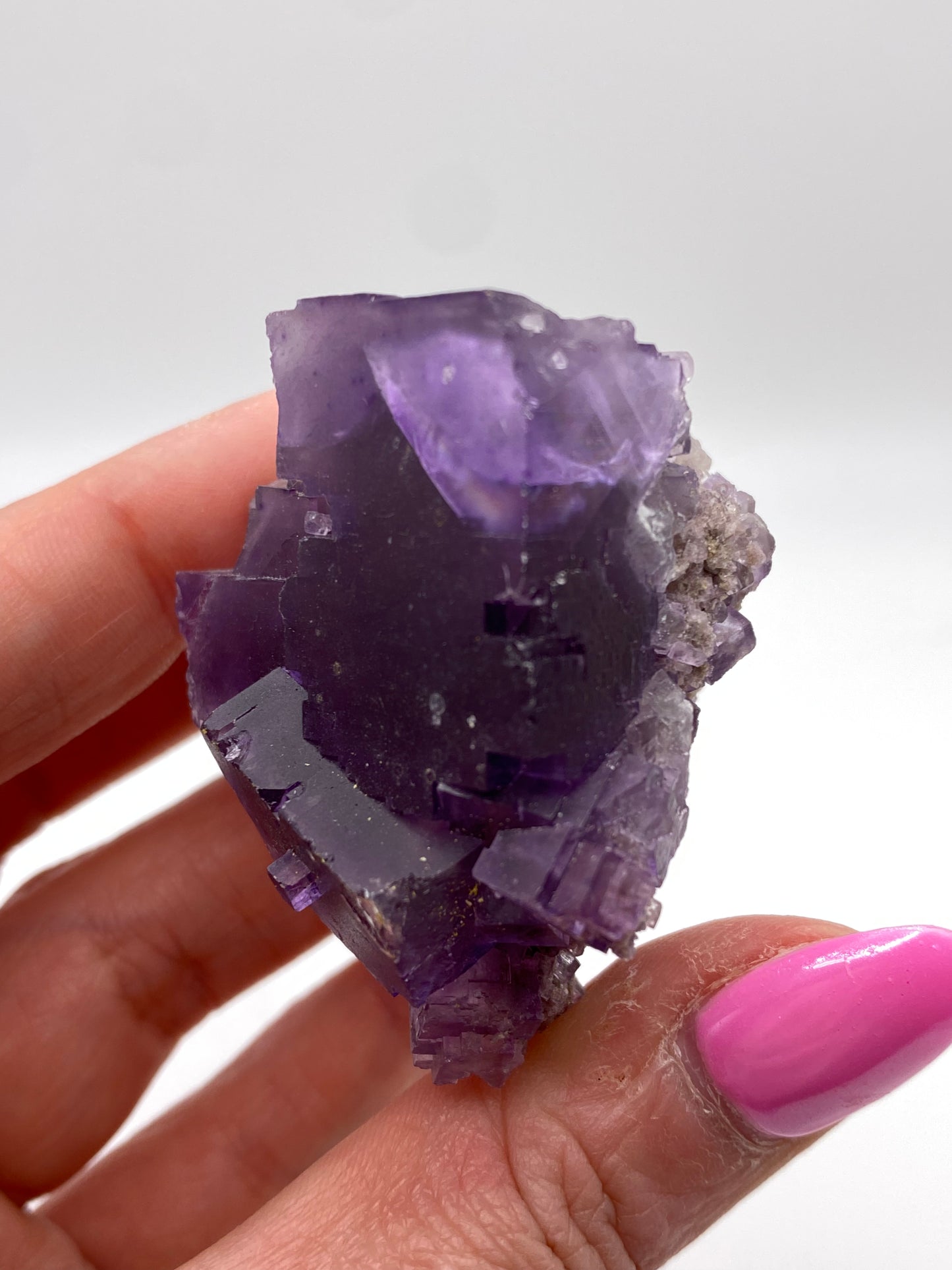 A hand with pink-painted nails holding a rough, translucent purple crystal against a plain white background. The jagged edges and irregular shape of the violet partial cube are evocative of The Crystalary's Fluorite, ex. Ben Clement- Cave in Rock, Hardin County, Illinois collection.