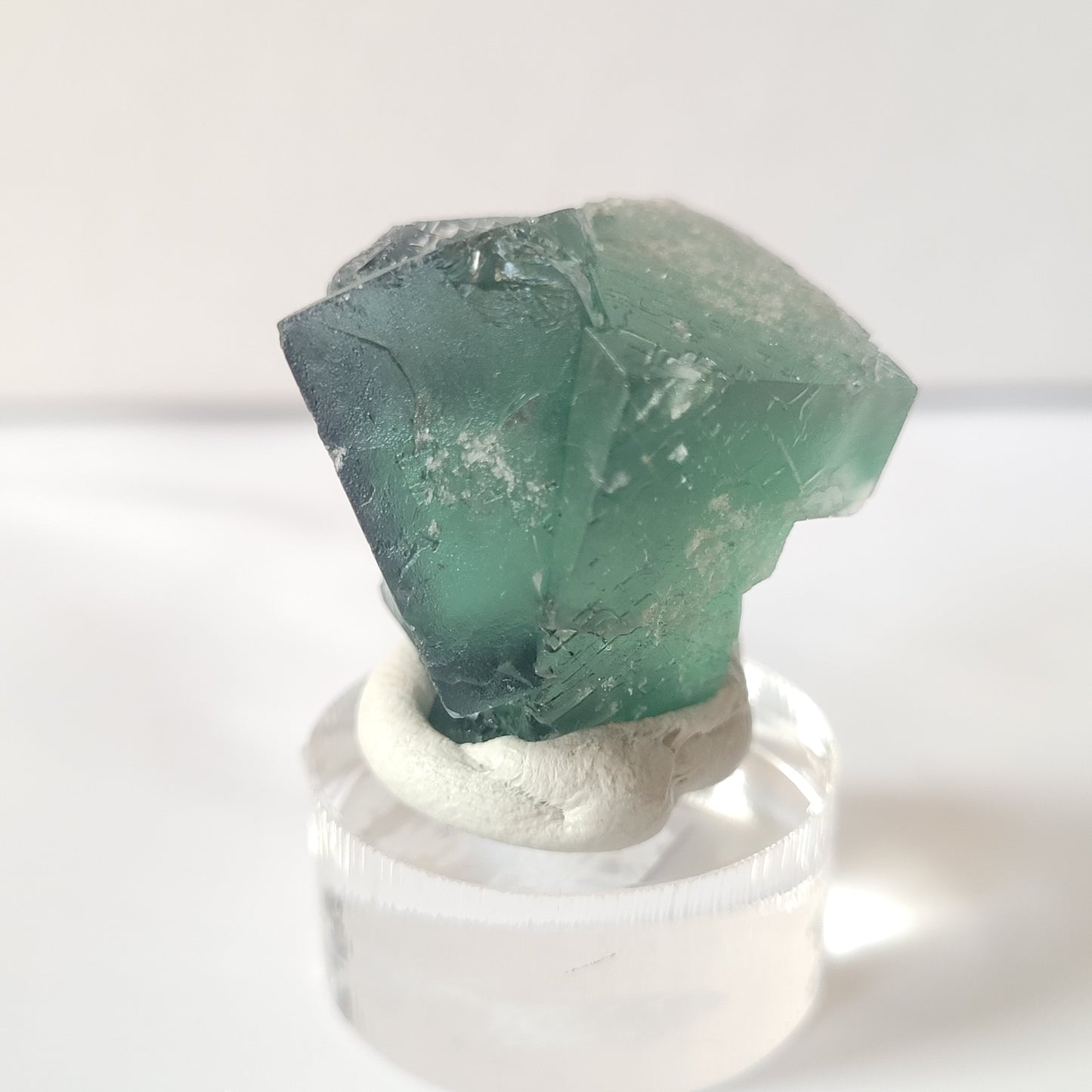 The Fluorite-Excavator Terminator from The Crystalary, sourced from Lady Annabella in Co. Durham, England, is a small translucent green crystal with a rough, angular shape. Mounted securely on a clear display stand using white putty and weighing 19 grams, this mini specimen stands out against the plain white background, showcasing its natural texture and color variations.