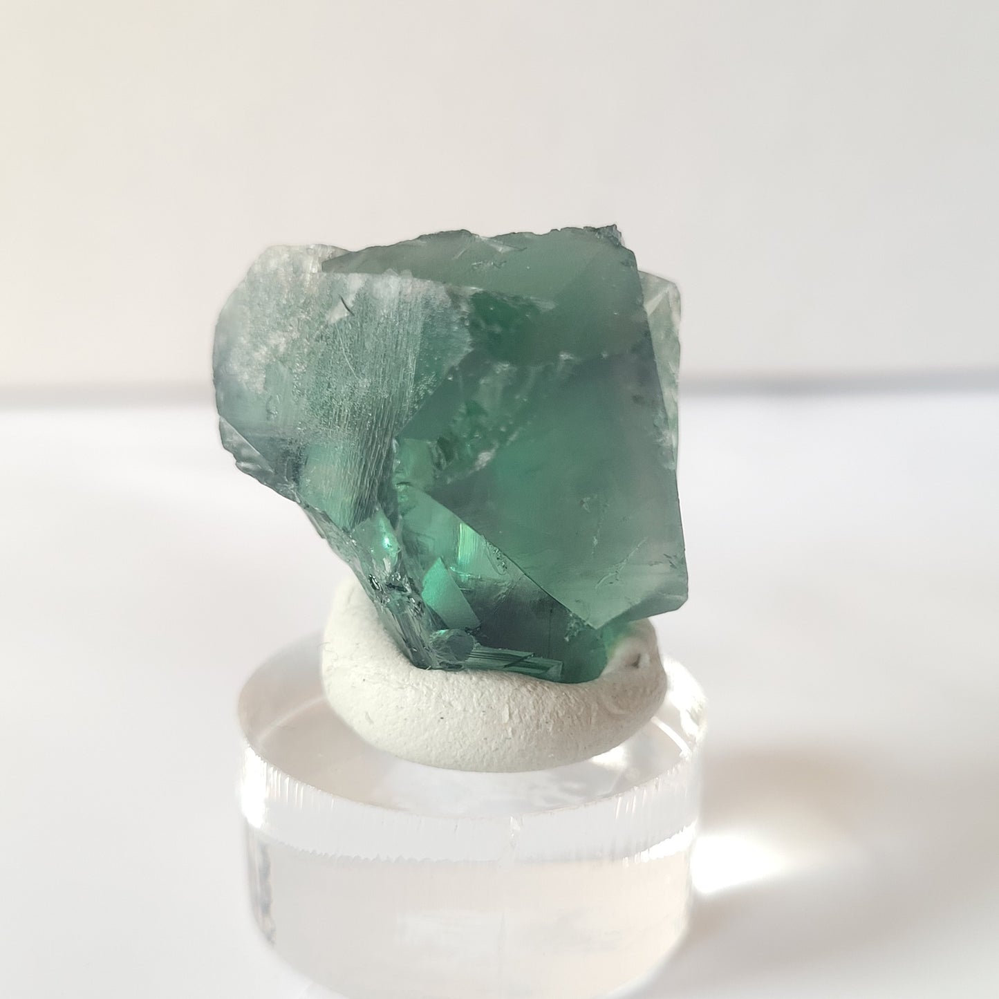 A close-up photo of "Fluorite- Excavator Terminator, Lady Annabella, Co. Durham, England" by The Crystalary, a green fluorite crystal chunk weighing 19 grams, mounted on a clear circular display stand with white adhesive. The background is plain and out of focus.