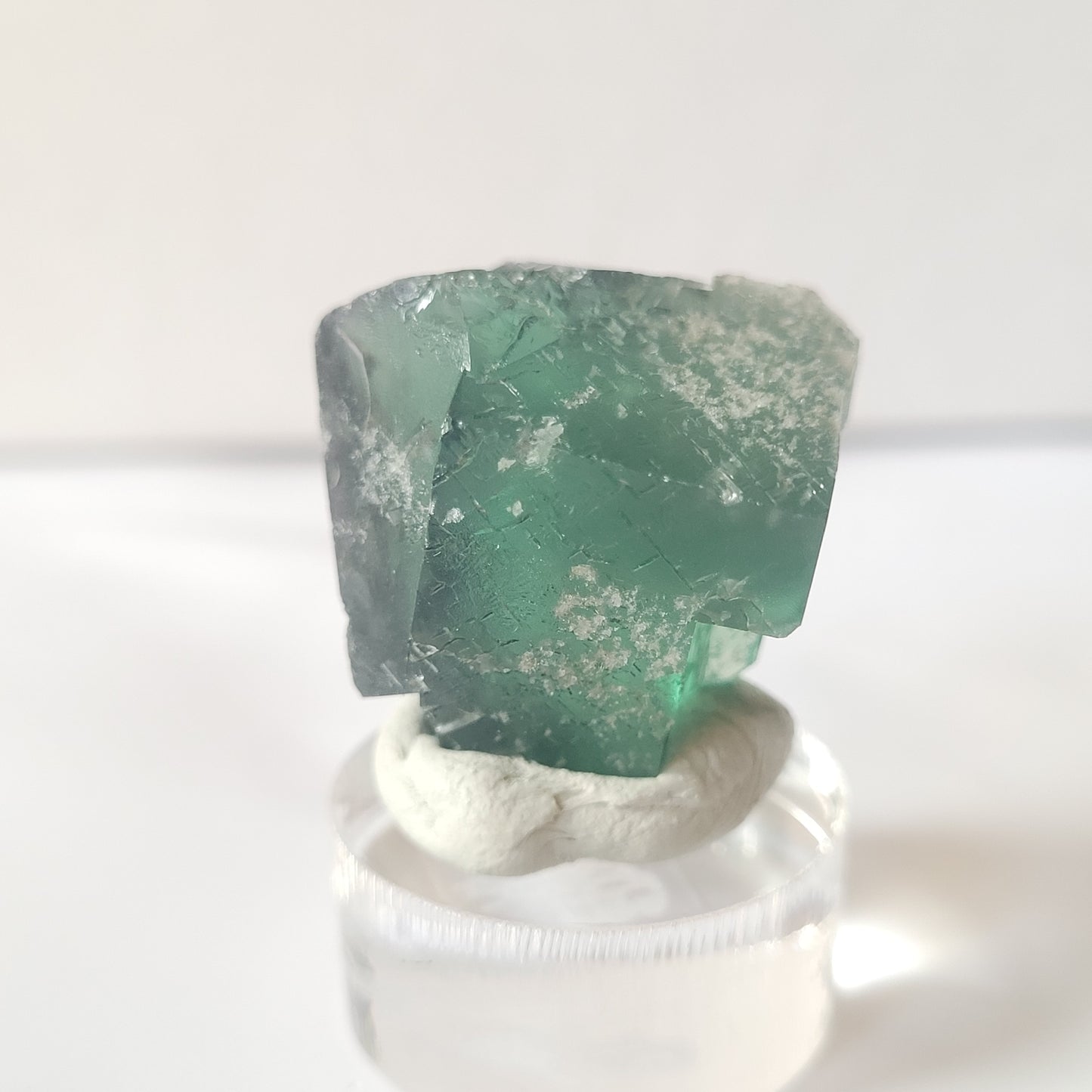 A close-up photograph showcases the Fluorite- Excavator Terminator, Lady Annabella, Co. Durham, England mini specimen from The Crystalary. The green fluorite crystal weighs 19 grams and exhibits translucent properties with cloudy white inclusions. It is elegantly positioned on a small round display stand crafted from a white clay-like material. The out-of-focus background features a light, neutral color palette.
