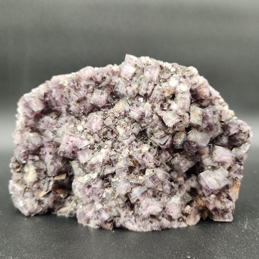 A cluster of translucent, purple cubic crystals forms a rugged mineral specimen. The background is a solid dark grey, which highlights the intricate details and various shades of light purple within the crystal formation, originating from the Fluorite, Funky February pocket in Greenlaws Mine, Co. Durham by The Crystalary.