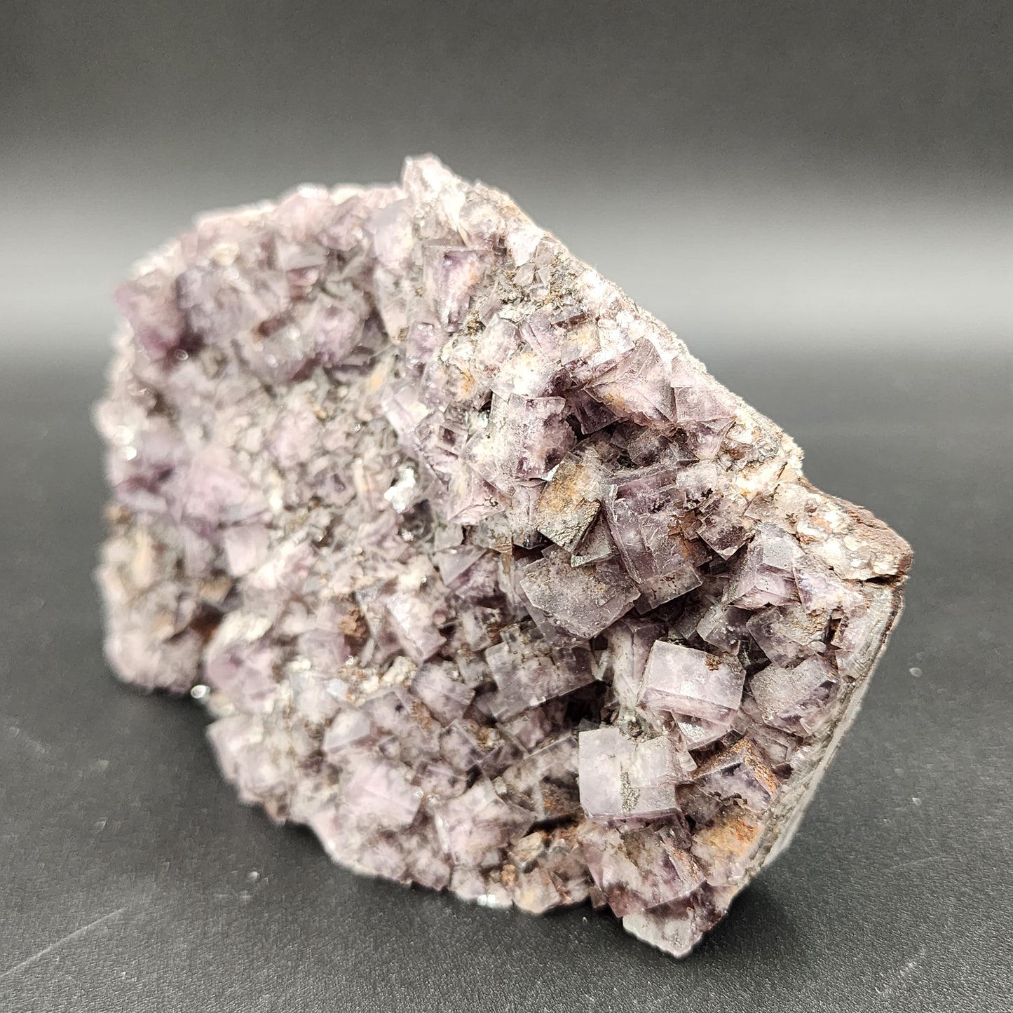 Displayed against a dark gray background, The Crystalary's large cluster of "Fluorite, Funky February" from Greenlaws Mine in Co. Durham, England showcases rough, uneven surfaces with cubic crystals varying in size. The semi-translucent fluorite crystals exhibit a subtle pastel hue.