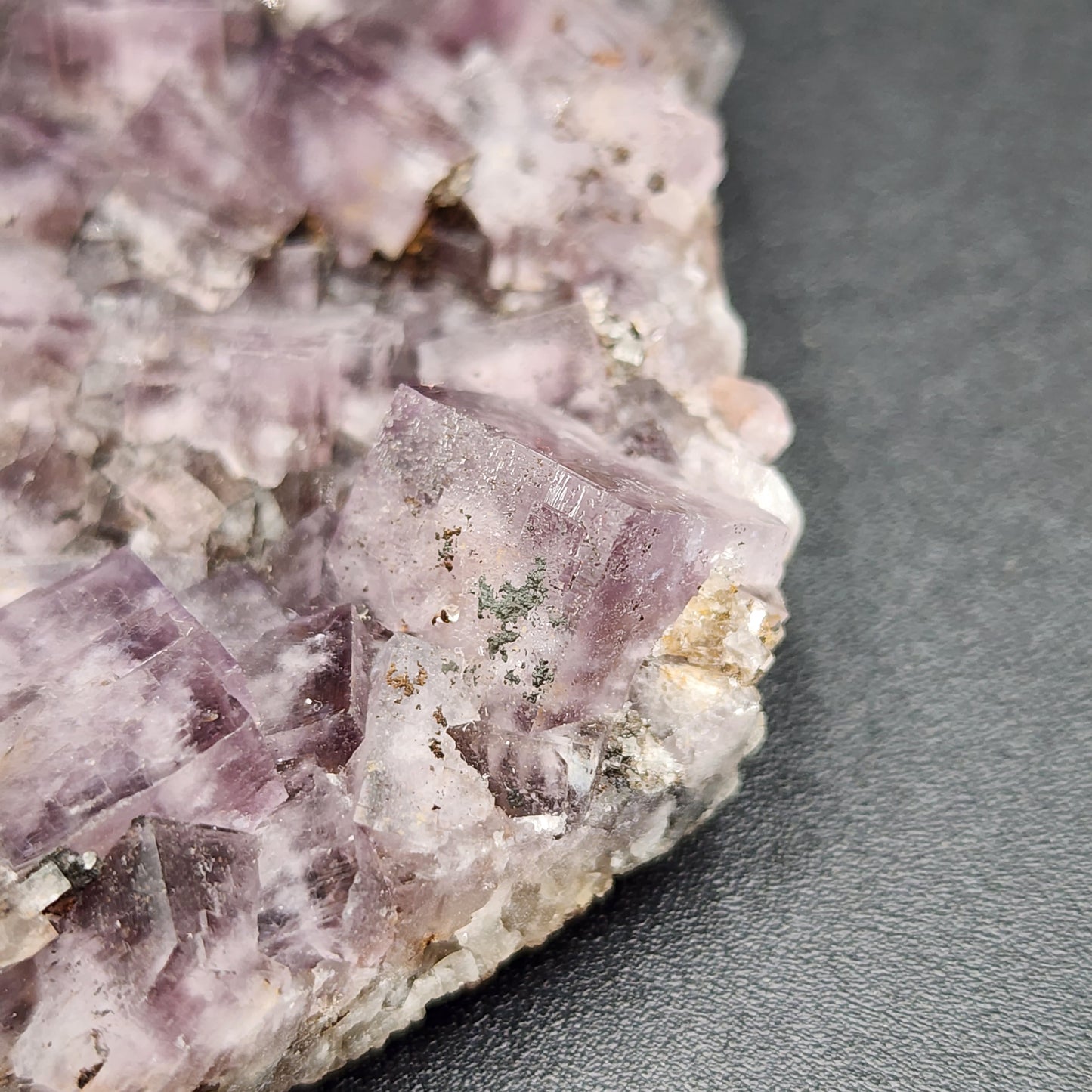 Close-up of a mineral specimen from Greenlaws Mine, Co. Durham, England, showcasing cubic, light purple crystals on a dark background. The crystals have a semi-transparent appearance with visible internal structures and fine details. This stunning piece is part of the "Funky February" collection by The Crystalary and is known as Fluorite.