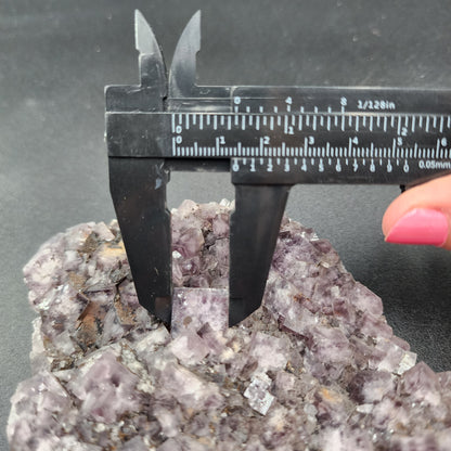 A hand with pink nail polish uses a black caliper to measure the dimensions of a rough, jagged purple crystalline rock specimen from Greenlaws Mine, Co. Durham. The rock's numerous small crystal formations perfectly capture the essence of The Crystalary's "Fluorite, Funky February" against the dark background.