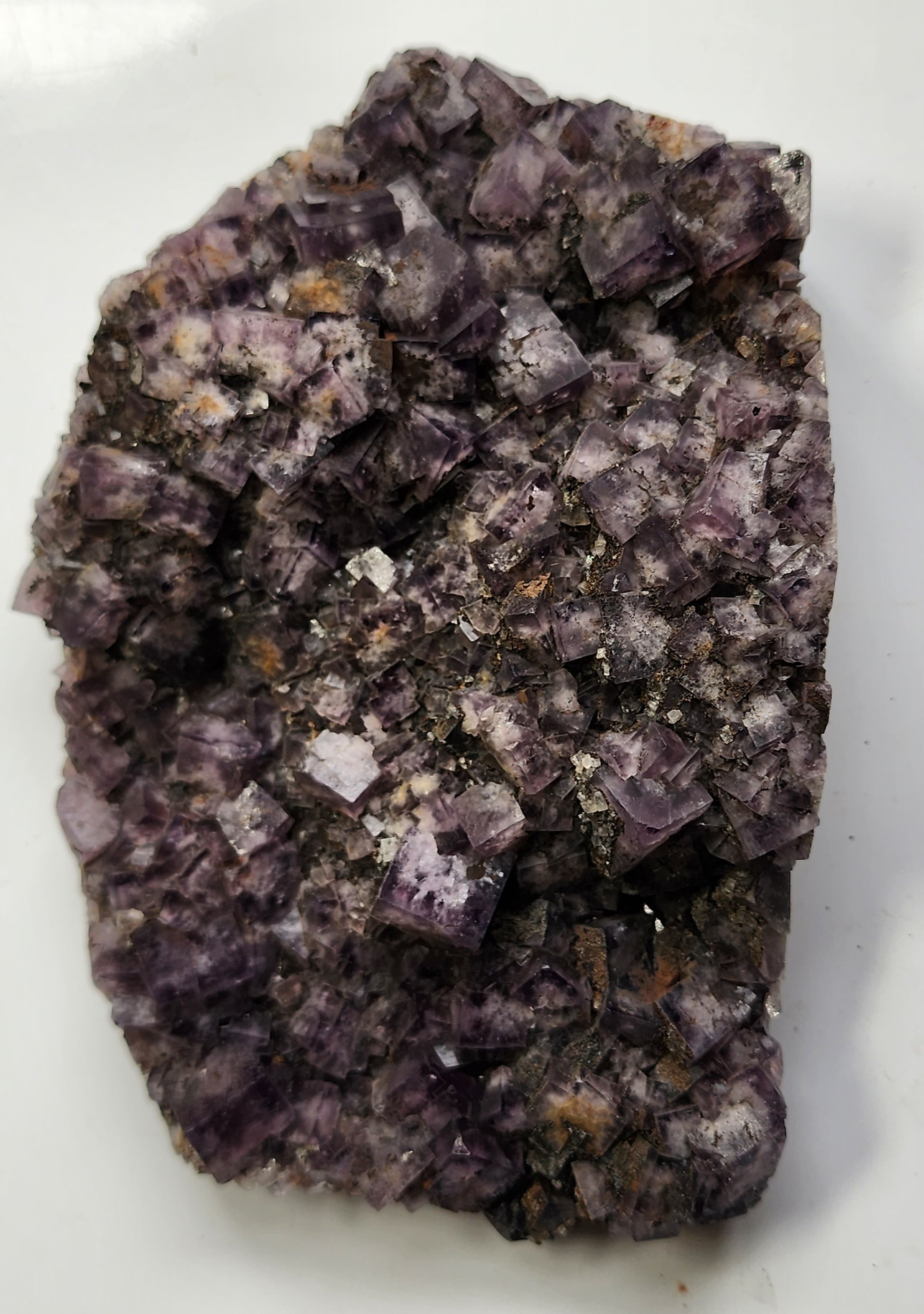 A close-up image of The Crystalary's Fluorite from the Funky February pocket at Greenlaws Mine, Co. Durham, England, showcasing a cluster of square and cubic formations with varying shades of purple and brown crystals on a white background. Some areas exhibit a slight shimmer due to light reflection.