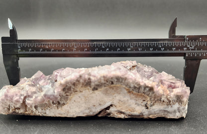 A close-up of a caliper measuring a piece of the rugged, crystalline Fluorite from The Crystalary's Funky February collection, sourced from Greenlaws Mine in Co. Durham, England. The rock showcases purple and gray hues and measures approximately 17.5 centimeters in length. The background is plain and dark, highlighting the distinct features of this unique specimen.