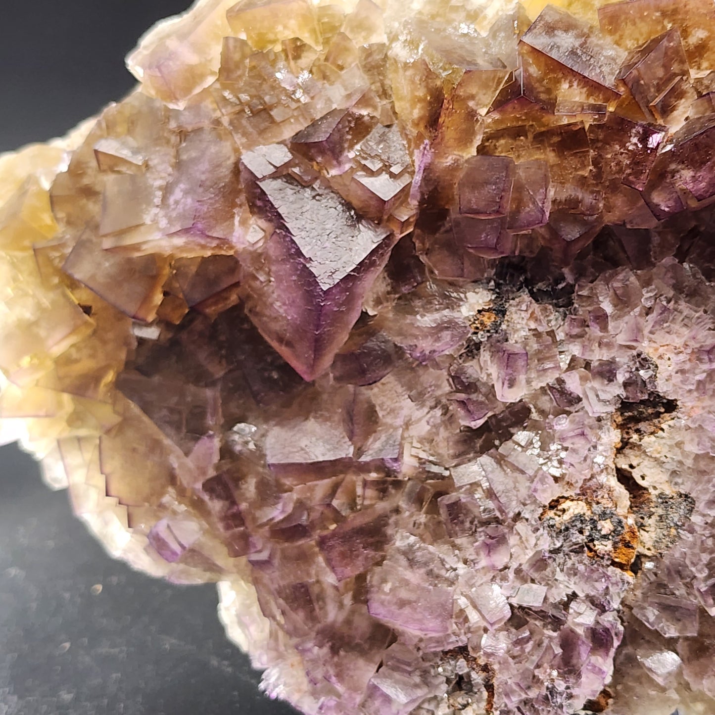 Close-up of a large Fluorite cluster with light and dark purple hues from The Crystalary. This stunning specimen, sourced from the Funky February Pocket at Greenlaws Mine in Co. Durham, exhibits sharp geometric formations and is partially embedded in a rock matrix. The surface appears translucent and glossy with intricate details and textures.