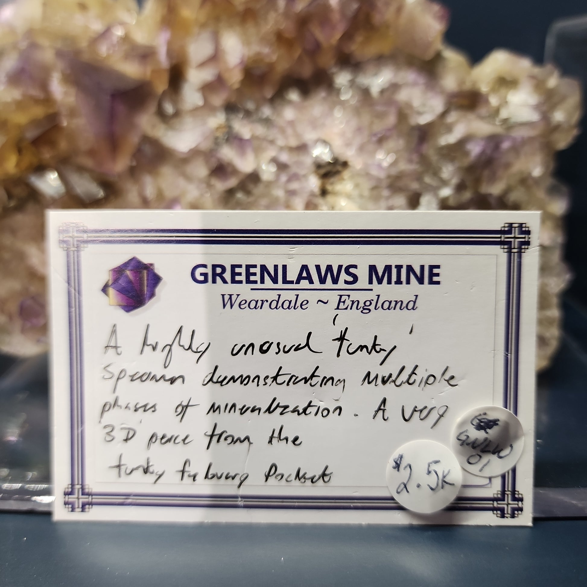 A close-up of a display card in front of a mineral specimen. The card reads "Greenlaws Mine, Co. Durham, England. A highly unusual 'funk' specimen demonstrating multiple phases of mineralization with a vibrant yellow core. A UV 3D piece from the Funky February Pocket." The Crystalary price tag shows "$2.5k.