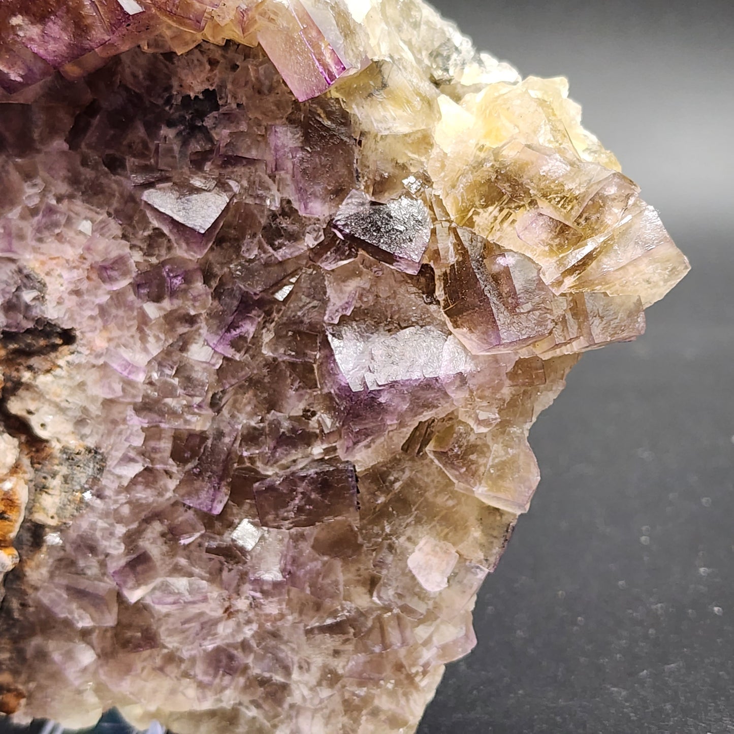 A close-up image of The Crystalary's Fluorite specimen from the Funky February Pocket of Greenlaws Mine in Co. Durham, England. This crystalline rock features clear and purple-hued formations with three-dimensional facets and jagged edges, creating a textured appearance. Light reflects off the varying surfaces of the mineral against a dark, plain background.
