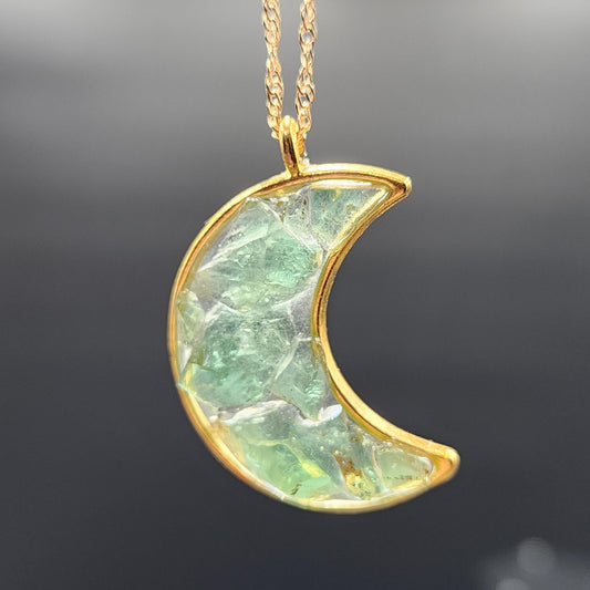 Introducing the Fluorite Gold Tone Moon Necklace from The Crystalary. This handmade pendant features a crescent moon with a gold frame, filled with green, translucent Rogerley Mine fluorite, and elegantly suspended from a delicate gold chain. The background is dark and slightly blurred for added emphasis.