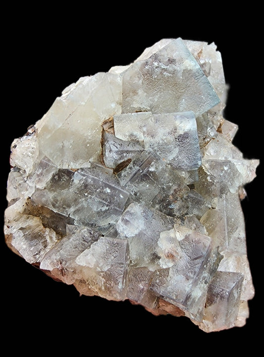 A close-up image of a cluster of clear and translucent cubic Fluorite crystals from The Crystalary's Green Toad Pocket at the Lady Annabella mine, set against a black background. The crystals exhibit natural formations with shades of light grey and subtle hints of purple, featuring irregular surfaces and mixed clarity.