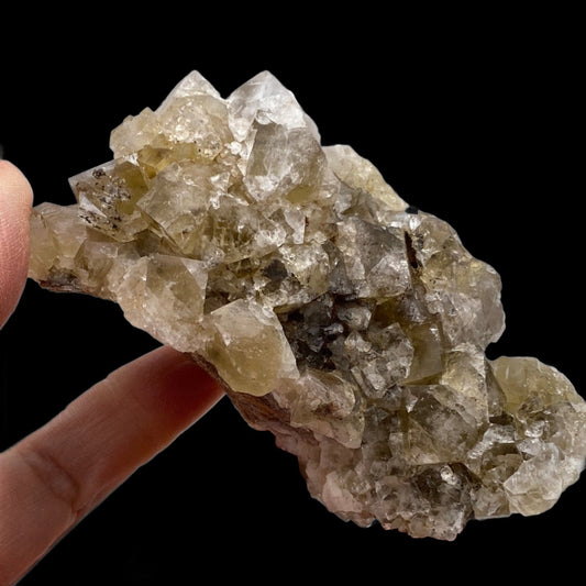 A hand holds a cluster of translucent crystals from The Crystalary's "Fluorite - Harvest Pocket, Lady Annabella, County Durham, UK" collection against a black background. The irregularly shaped crystals vary in size and have a pale yellow-brown tint with subtle fluorite interspersed. The fingers are visible, giving an idea of the mineral’s size.