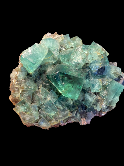 A cluster of translucent, cubic green fluorite crystals on a contrasting black background. Known for their UV reactivity, these crystals from The Crystalary's Fluorite- Heavy Metal Pocket collection, sourced from the Diana Maria Mine in Weardale, Co Durham, UK, exhibit varying shades of green with subtle hints of blue. Some edges reflect light, highlighting their intricate natural formation.