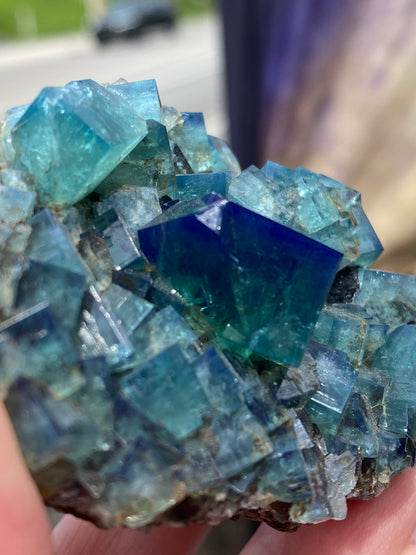 Close-up of a person holding a cluster of vibrant green Fluorite- Heavy Metal Pocket crystals from the Diana Maria Mine in Weardale, Co Durham, UK. These crystals, offered by The Crystalary, exhibit a cubic structure and glisten in natural light, showcasing their colorful and intricate patterns. A blurred background suggests an outdoor setting.