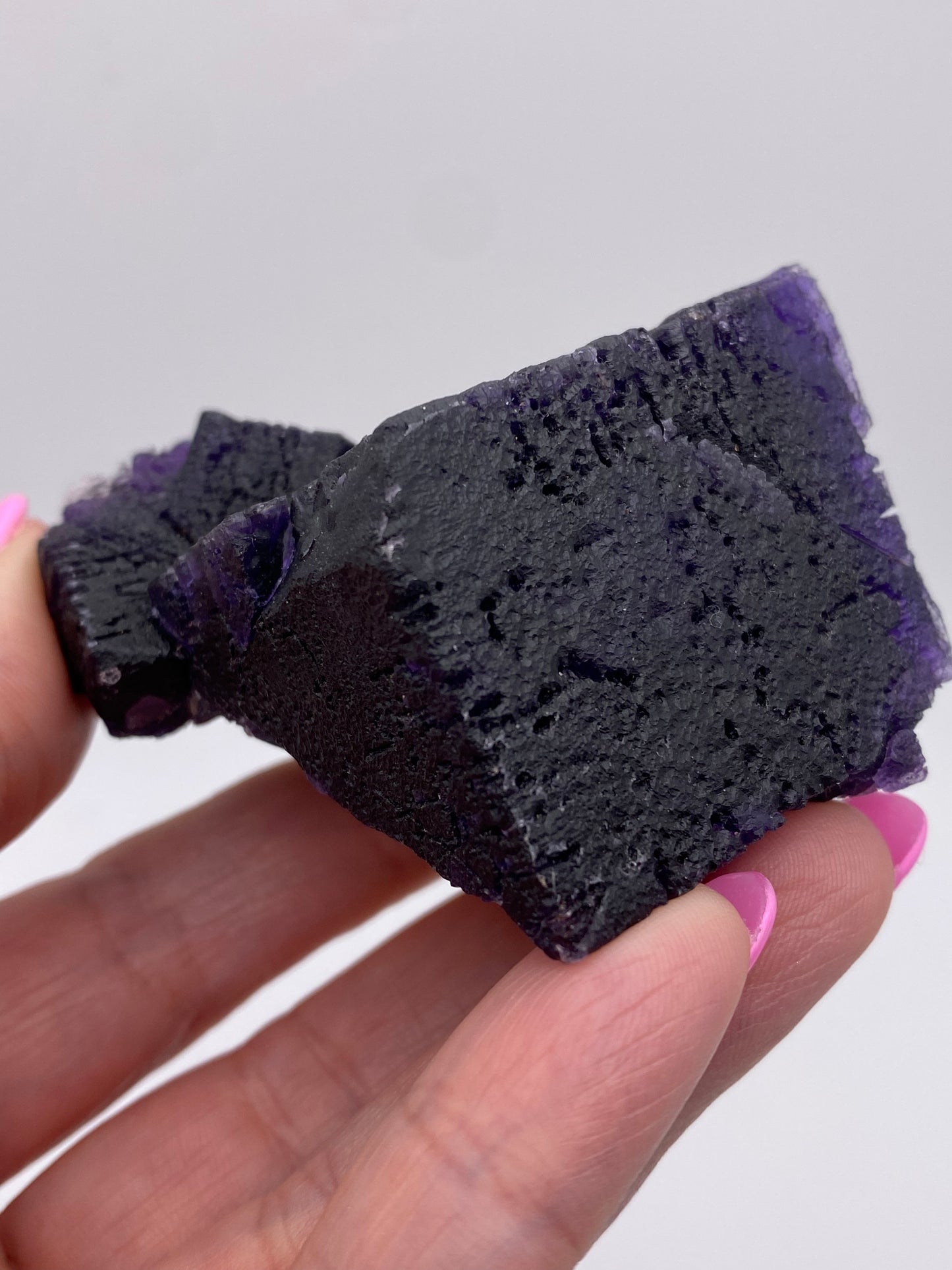 Close-up image of a hand holding The Crystalary's Fluorite from Hill-Ledford Mine in Cave-In-Rock, Hardin County, Illinois, USA. The dark purple crystal features cubic formations with a rough texture. The person holding it sports pink nail polish, while the background is blurred and light gray.