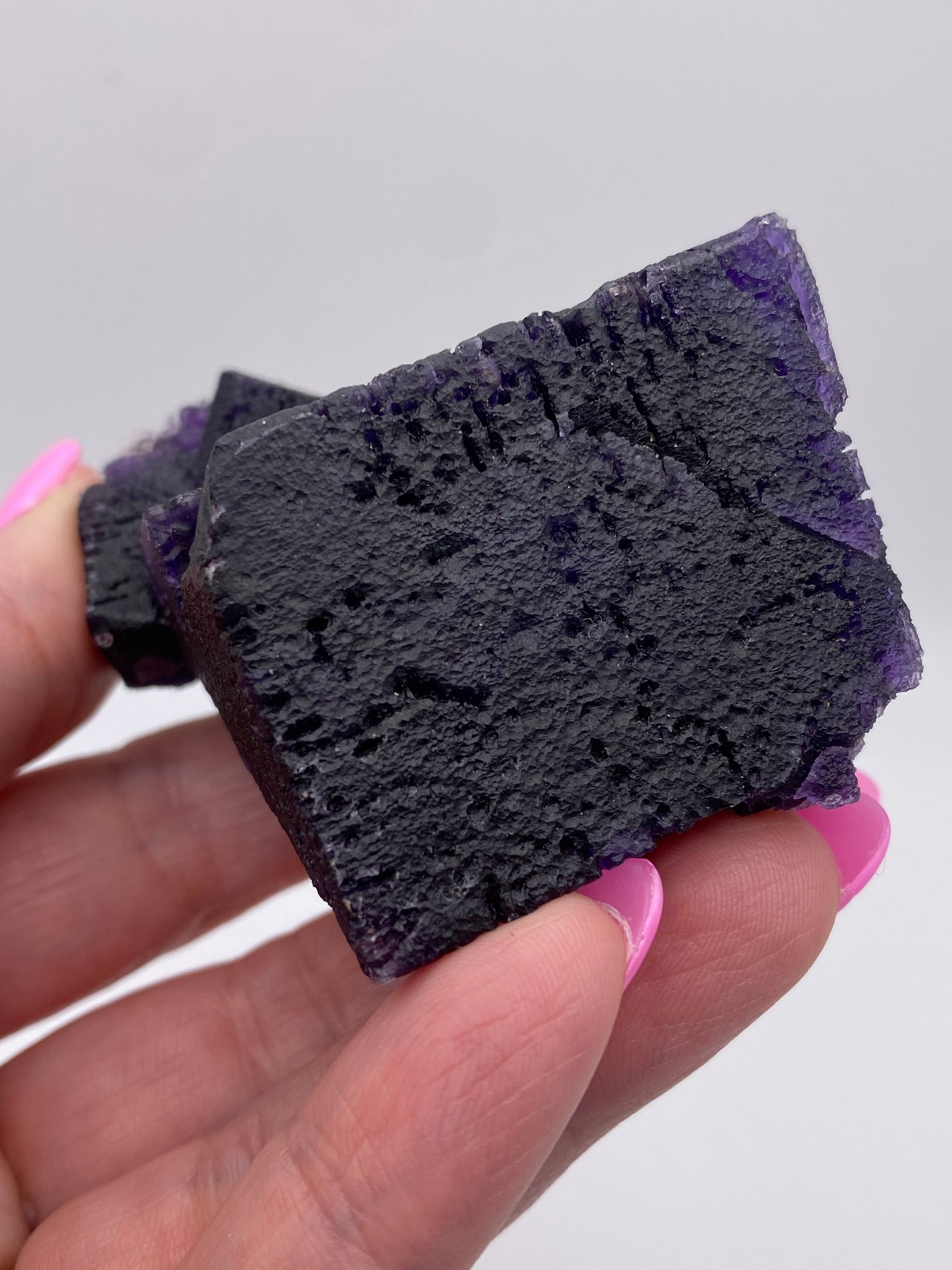 A hand with pink nail polish holds a stunning, dark purple Fluorite from The Crystalary, sourced from the Hill-Ledford Mine in Cave-In-Rock, Hardin County, Illinois against a plain background. The cube-shaped crystal features a rough and textured appearance reminiscent of the renowned deposits in the area.