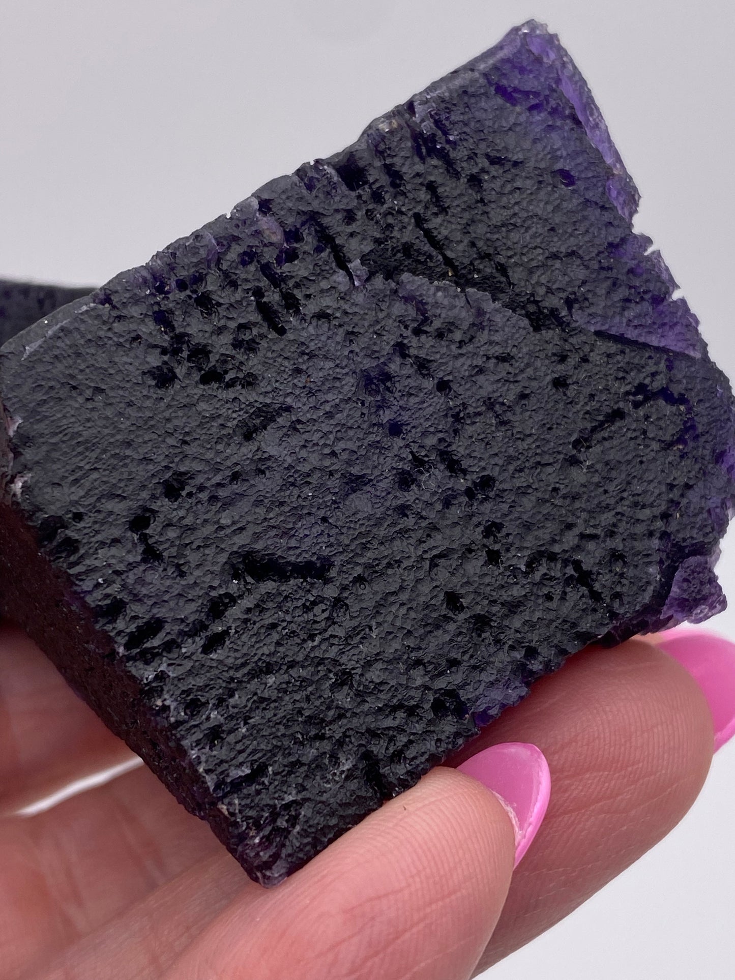 A close-up image of a person holding a dark purple, rough-textured The Crystalary's Fluorite from the Hill-Ledford Mine in Cave-In-Rock, Hardin County, Illinois, USA with their fingers. The person's nails are painted bright pink. The background is plain and out of focus, emphasizing the mineral's details.