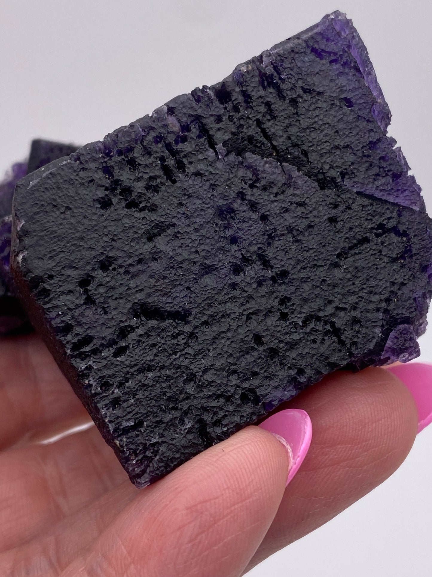 Close-up of a hand with pink-painted nails holding a dark purple, rough-textured mineral with a cubic structure. This Fluorite from The Crystalary, specifically sourced from the Hill-Ledford Mine in Cave-In-Rock, Hardin County, Illinois, USA features an uneven surface with streaks and a deep purple coloration. The background is plain and white.